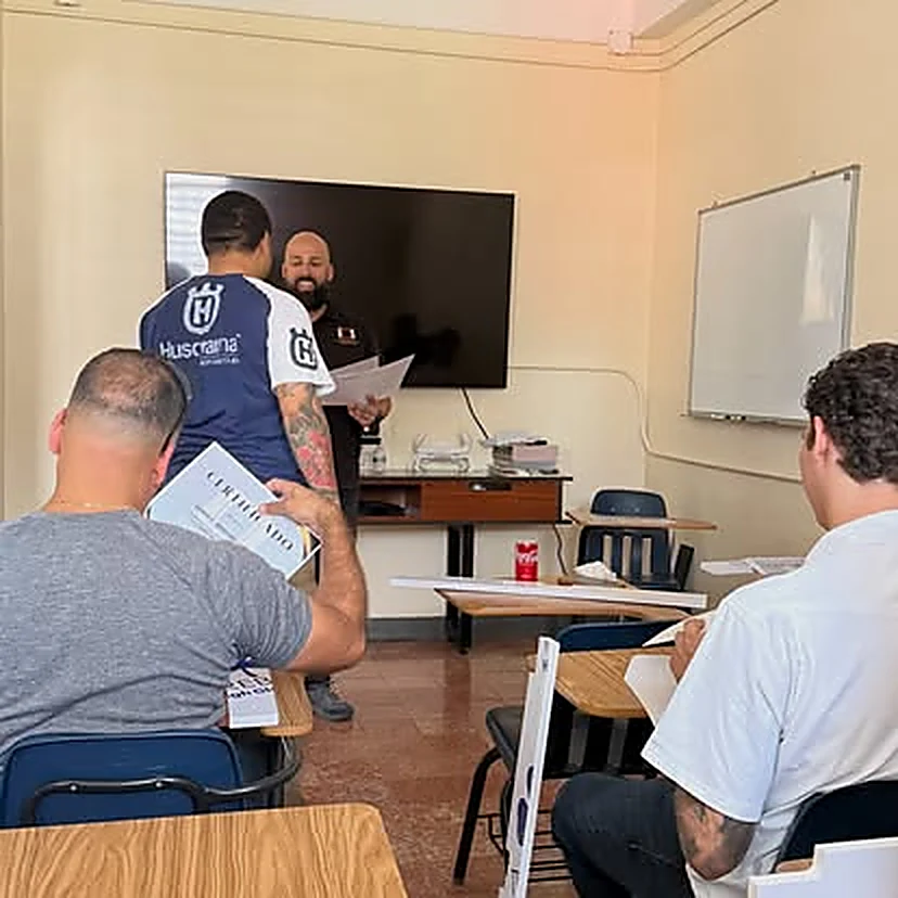 Salón de clase con el instructor al frente de una pantalla y estudiantes sentados tomando notas.